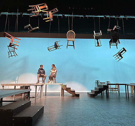 actors on stage, under hanging chairs