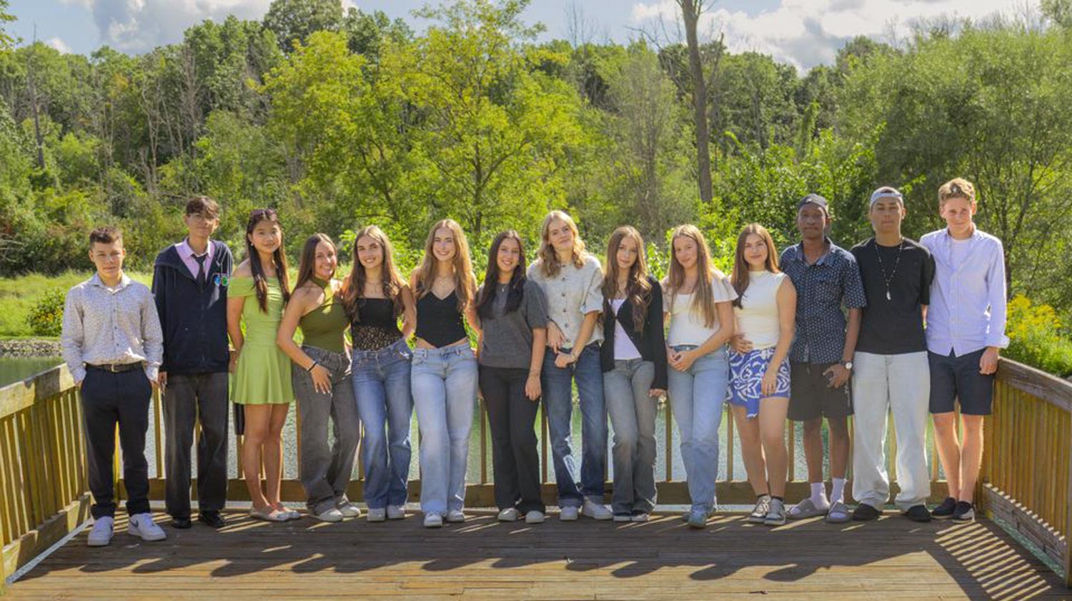 Group of 13 young people standing on a wooden deck with greenery in the background