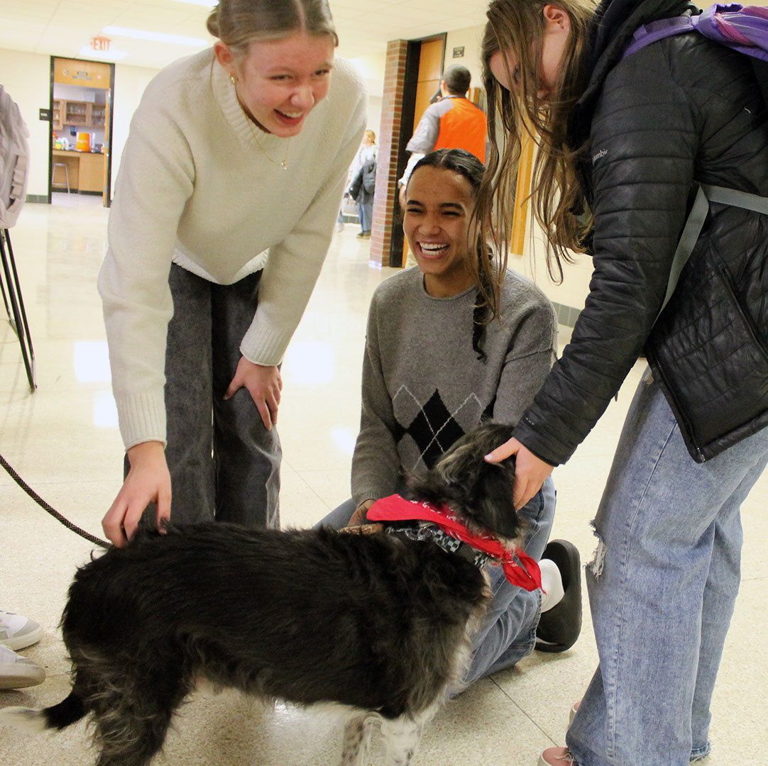 three students smiling while petting a dog in a PHS hallway