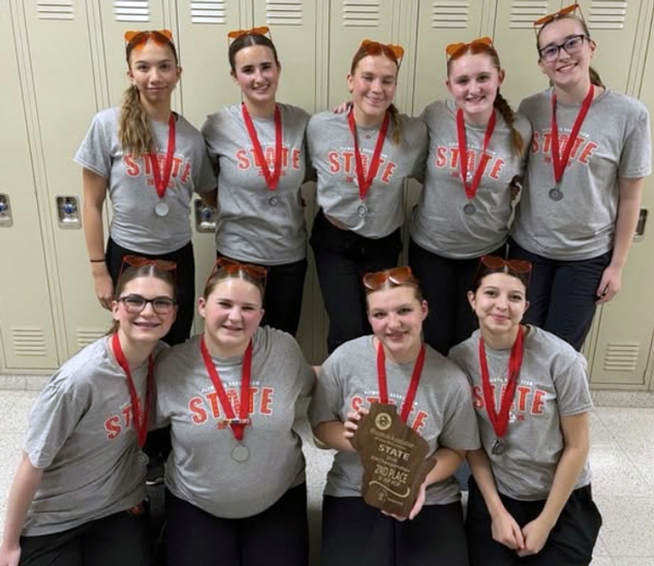 9 dancers wearing grey T-shirts and medals, posing with a wooden Wisconsin-shaped plaque